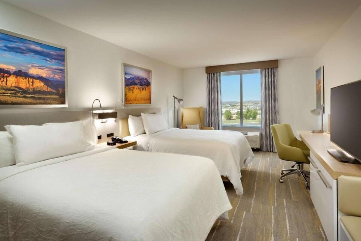 A hotel room with two beds, a desk with a TV, a chair, a lamp, and a window with a view.