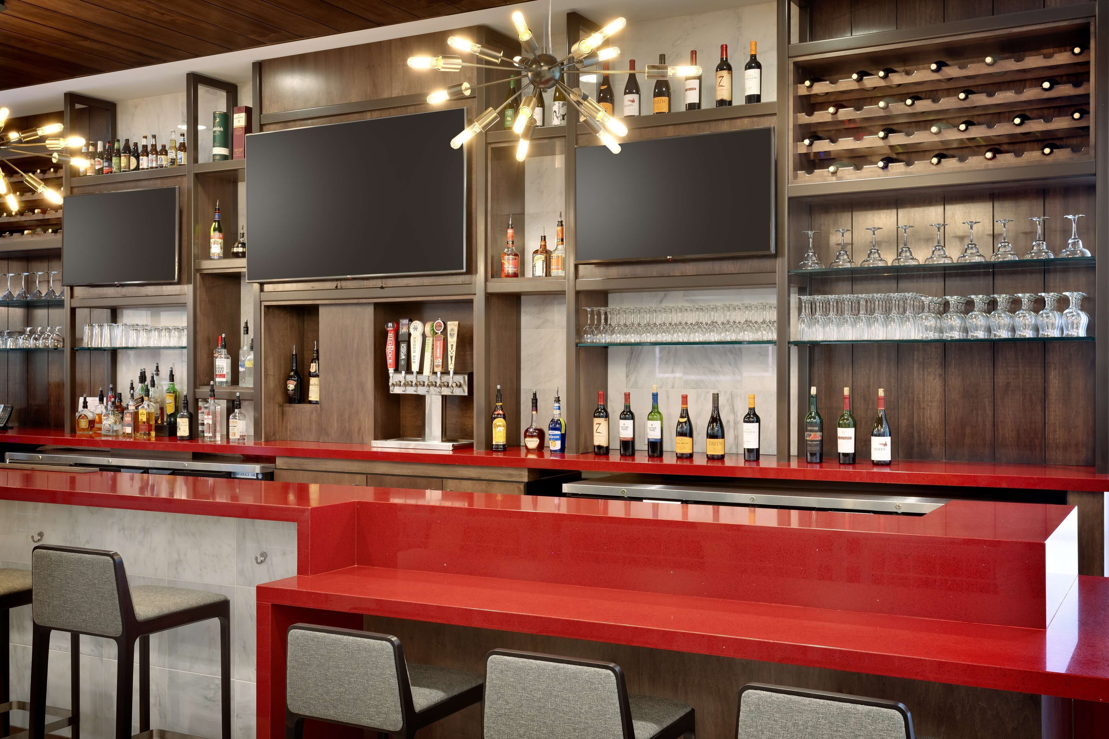 Una zona de bar con mostrador rojo, varias estanterías llenas de botellas y vasos, y taburetes.