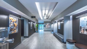 A modern hotel corridor with a long hallway, wall-mounted artwork, and a sign indicating the direction to the bathrooms. - Hampton Inn & Suites Tallahassee Capitol - University (Tallahassee)