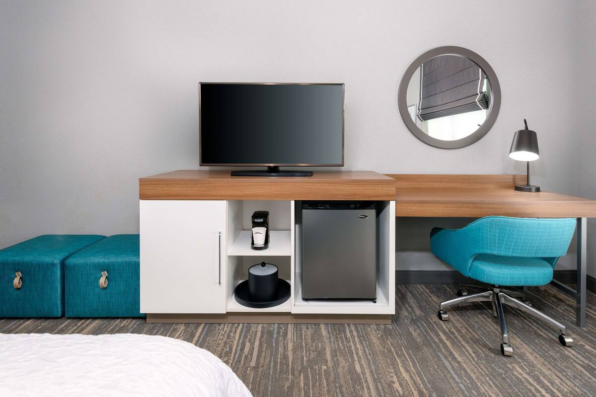 A hotel room with a wooden desk, a television, a mirror, a desk lamp, a refrigerator, a teal chair, and a bed with a white comforter.