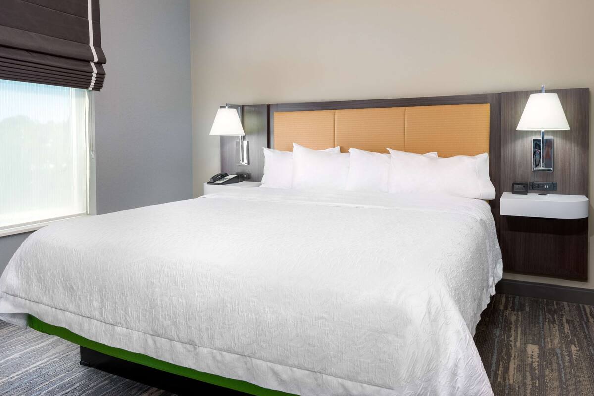 A hotel room with a large bed, two bedside tables with lamps, a nightstand, and a window with blinds.