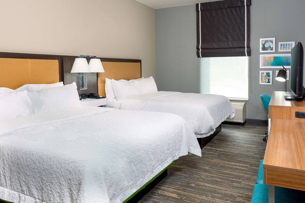 A hotel room with two beds, a desk, a chair, a TV, and a window with blinds.