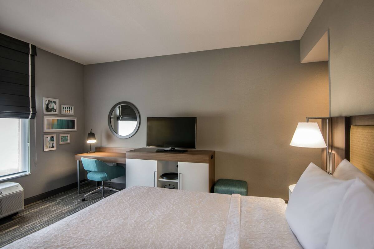 A hotel room with a bed, a desk with a TV, a chair, a lamp, and a window with blinds.