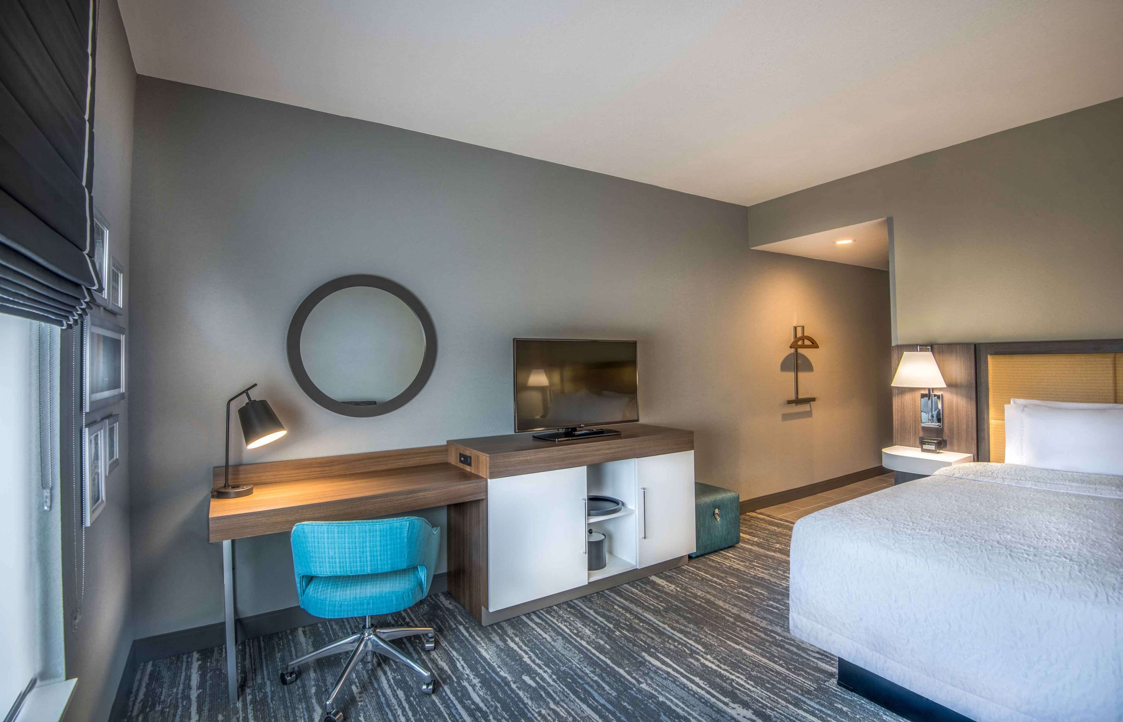 A hotel room with a bed, a desk with a chair, a round mirror, a television, and a bedside table with a lamp.