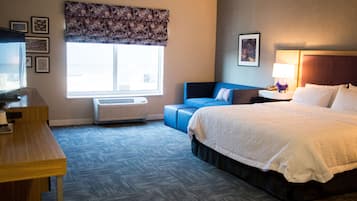 Habitación de hotel con cama, escritorio, silla, ventana con persianas, aire acondicionado y una pared con cuadros.