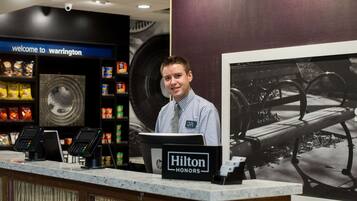 Ein Hotel-Rezeptionsbereich mit einem Schild, das „Hilton HONORS“ zeigt, und einem Schild mit der Aufschrift „Willkommen in Warrington“ über der Theke.