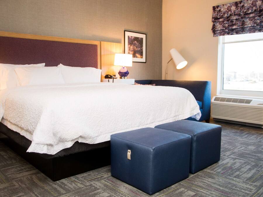 A hotel room with a large bed, a nightstand with a lamp, a chair, and a window with an air conditioner.