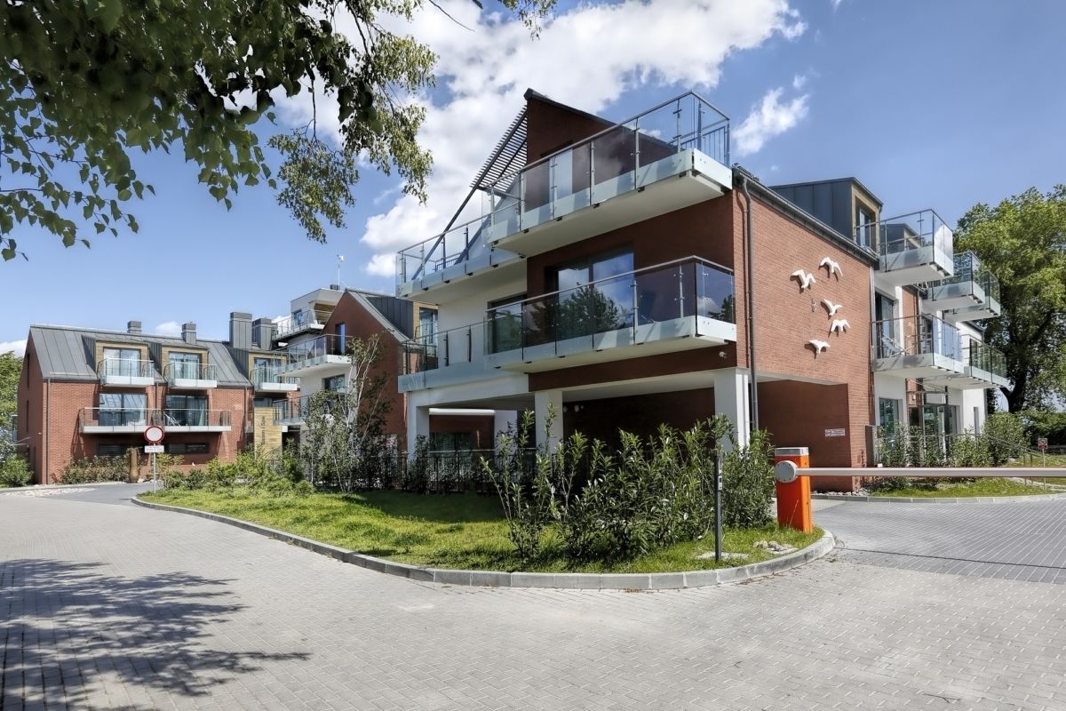 Um edifício residencial moderno de vários andares, com varandas, entrada gradeada e gramado bem cuidado.