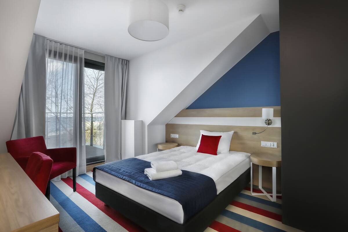 A modern hotel room with a bed, a desk, a chair, and a view of the outdoors.