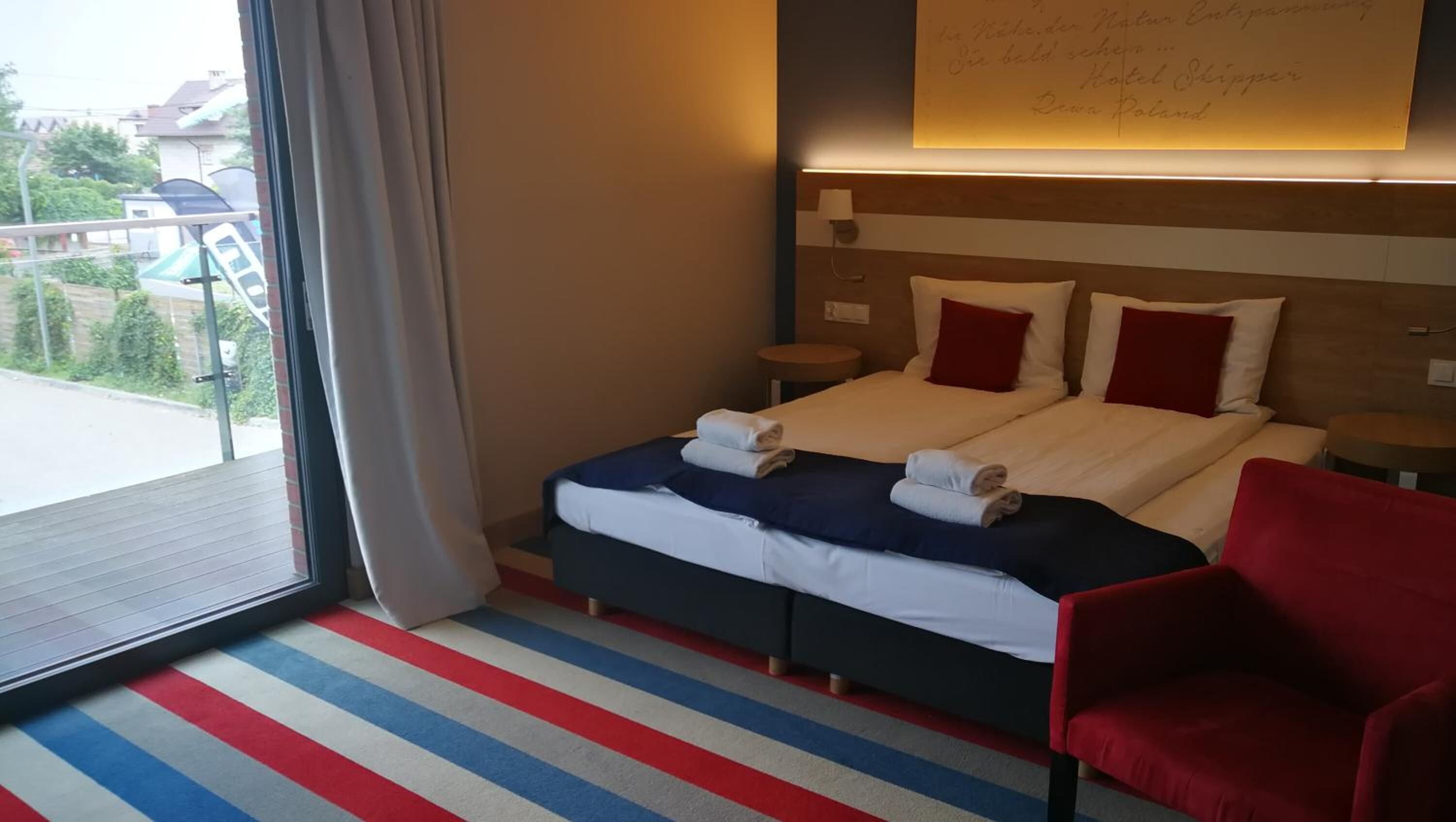 Een hotelkamer met een bed, twee nachtkastjes, een rode stoel, een gestreept tapijt en uitzicht naar buiten.