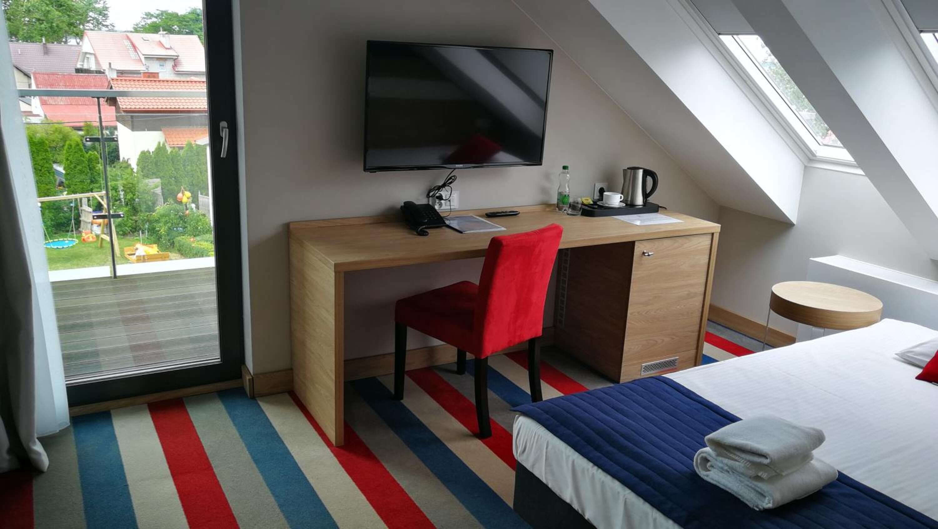 Una habitación de hotel moderna con un escritorio de madera, una silla roja, una cama con alfombra azul y blanca a rayas, un televisor montado en la pared y vistas al balcón exterior.