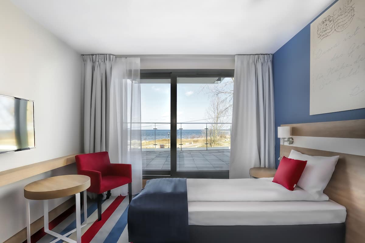 A hotel room with a bed, a red chair, a small table, and a large window offering a view of the beach.