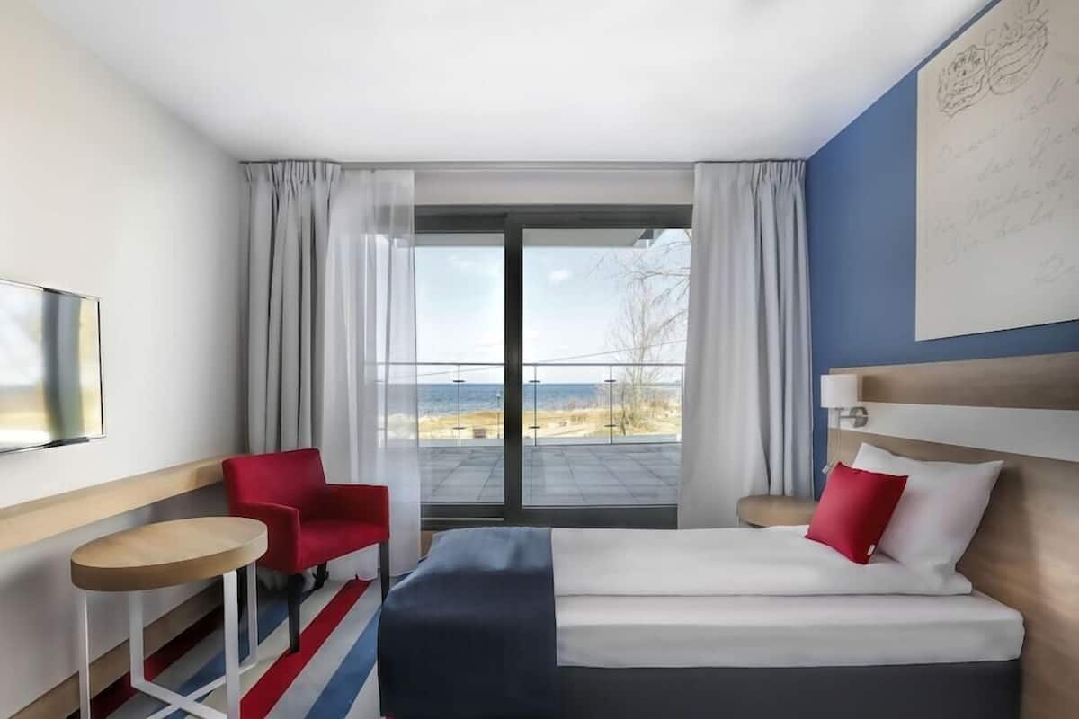 A hotel room with a bed, a red chair, a small table, and a view of the outdoors.
