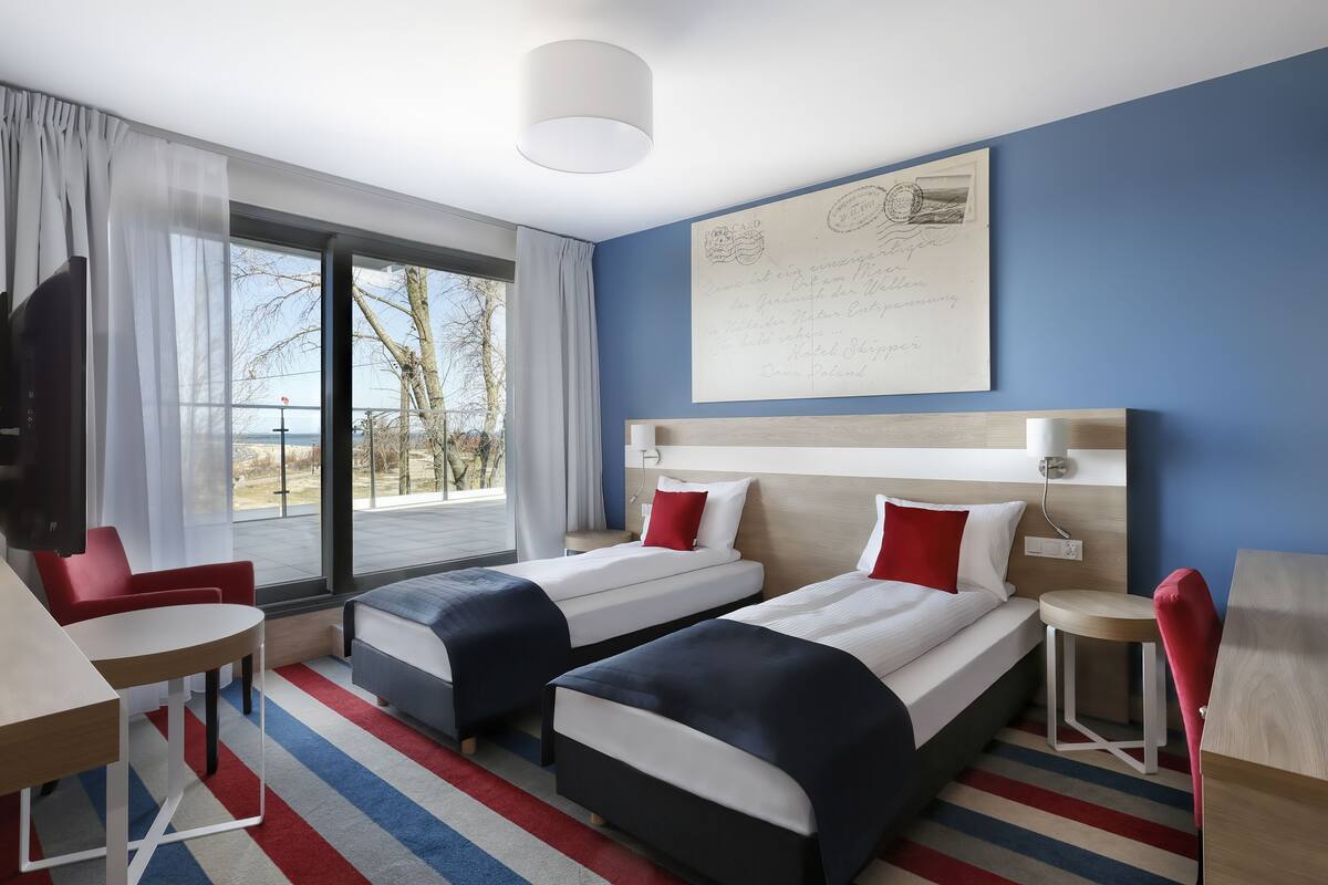 A hotel room with two beds, a desk, a chair, and a large window.