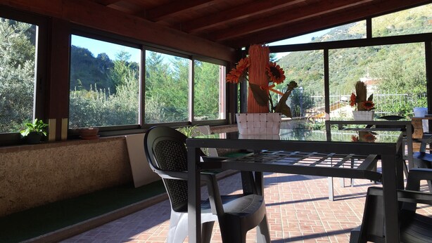Un patio techado con una mesa y sillas, ventanas amplias que dan a vistas de vegetación y colinas, y una lámpara colgante.