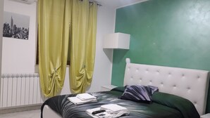 Habitación de hotel con cama, mesita de noche, lámpara, radiador y ventana con cortinas.