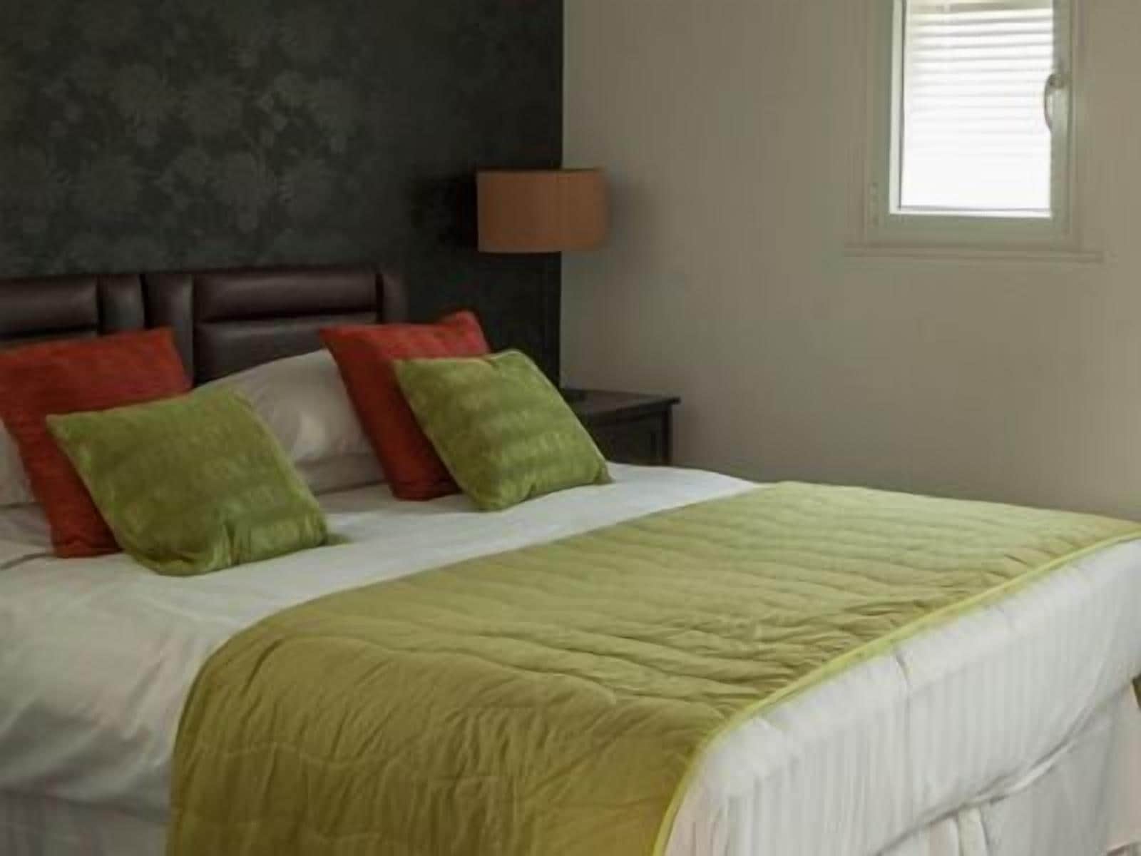 A bed with a green and white bedspread, two red and two green pillows, a dark headboard, a bedside lamp, and a window with blinds.
