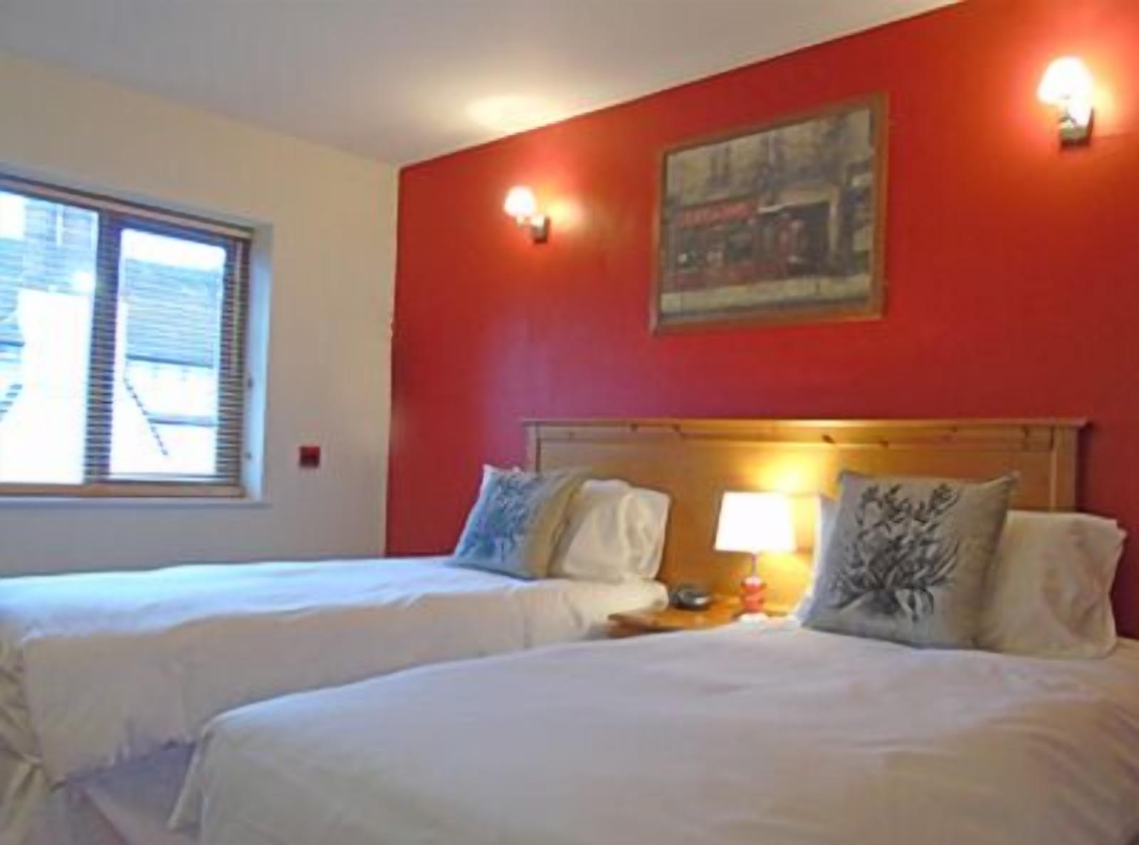 Een hotelkamer met twee bedden, een raam met zonwering, een schilderij aan de muur en een nachtkastje met een lamp.