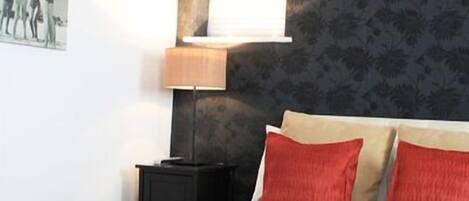 A bedroom with a bed, two red pillows, a nightstand, a lamp, and a wall with a floral pattern.