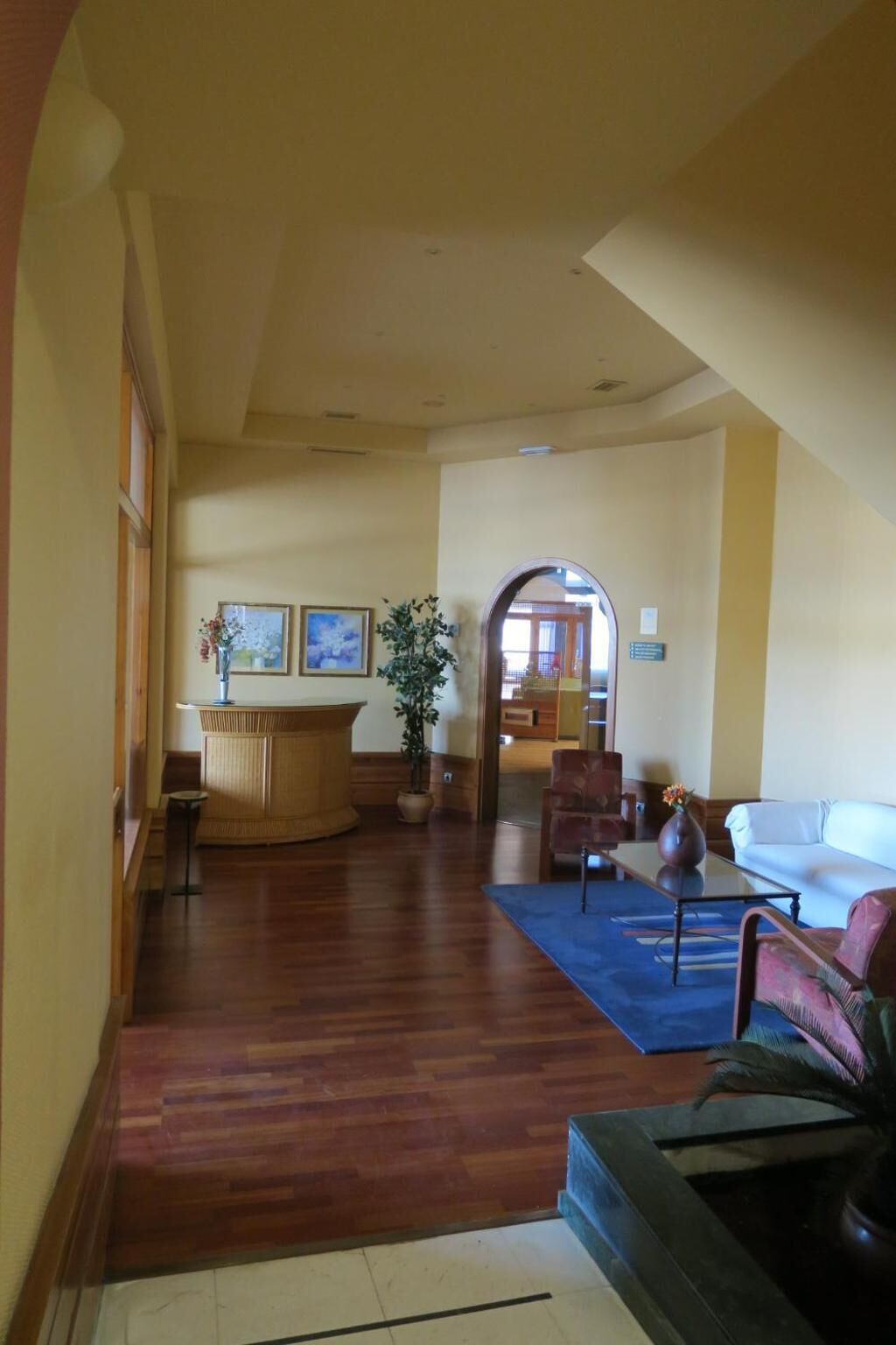 Un vestíbulo amplio con suelo de madera, un mostrador de recepción, una zona de estar con sofá y sillones, y una planta grande.