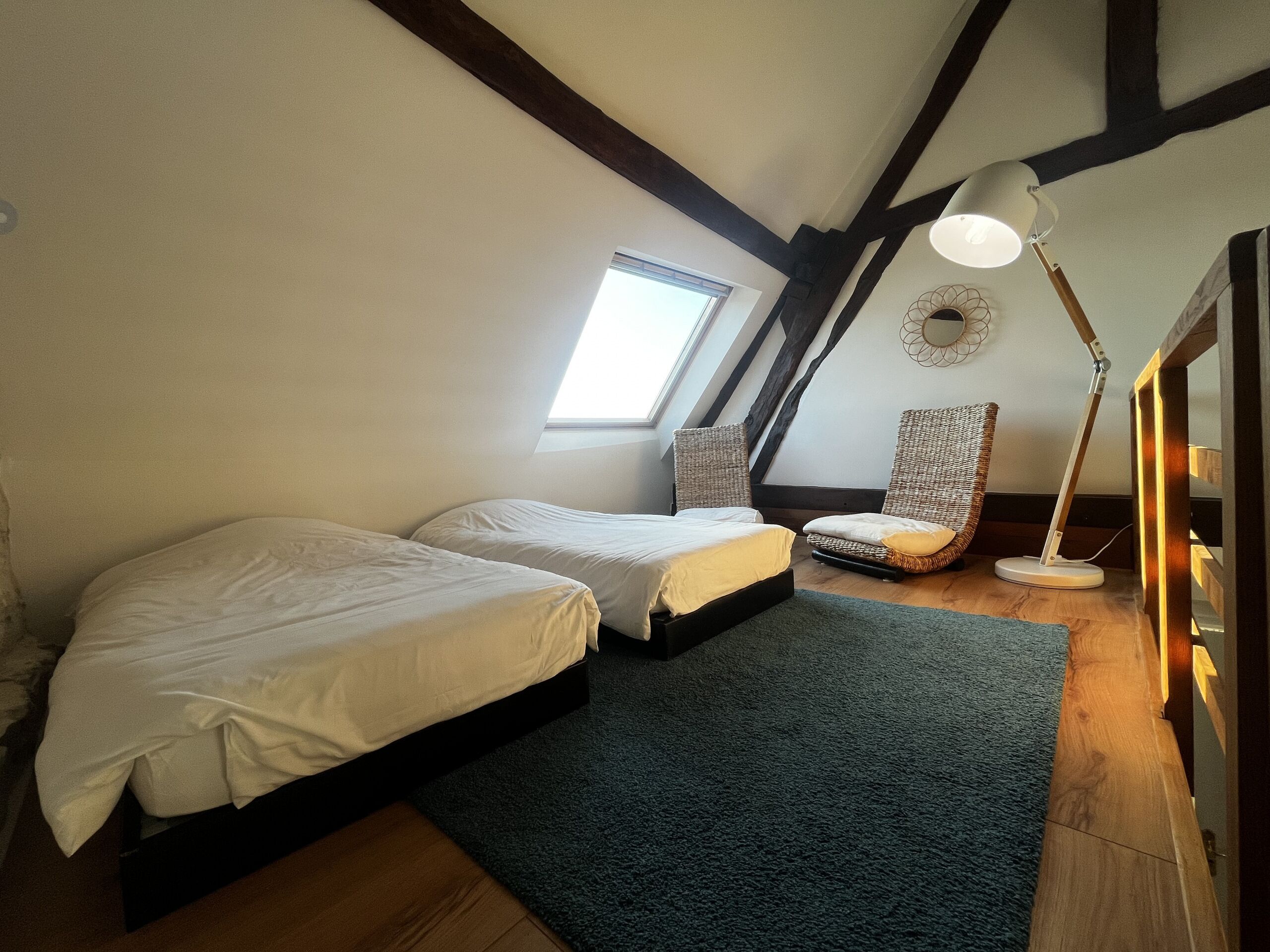 Una habitación con dos camas, una lámpara de pie, una silla de mimbre y una ventana.
