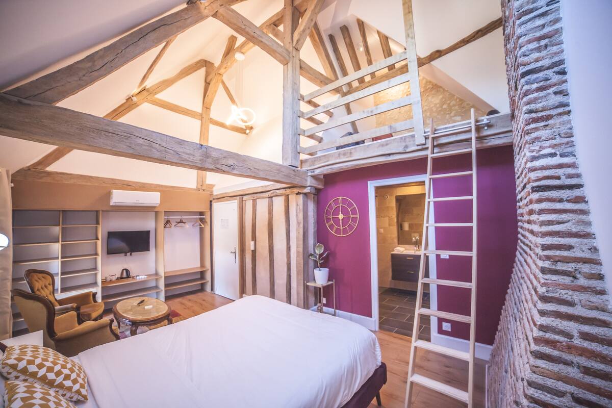 A bedroom with a large bed, wooden beams, a TV, a chair, a small table, and a bathroom with a red wall and a ladder.