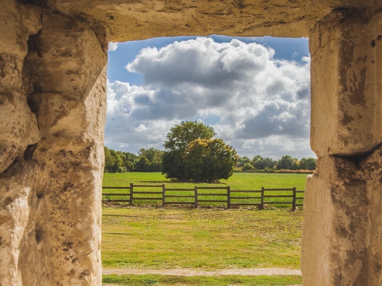 A stone archway framing a view of a grassy field with a wooden fence and a tree.