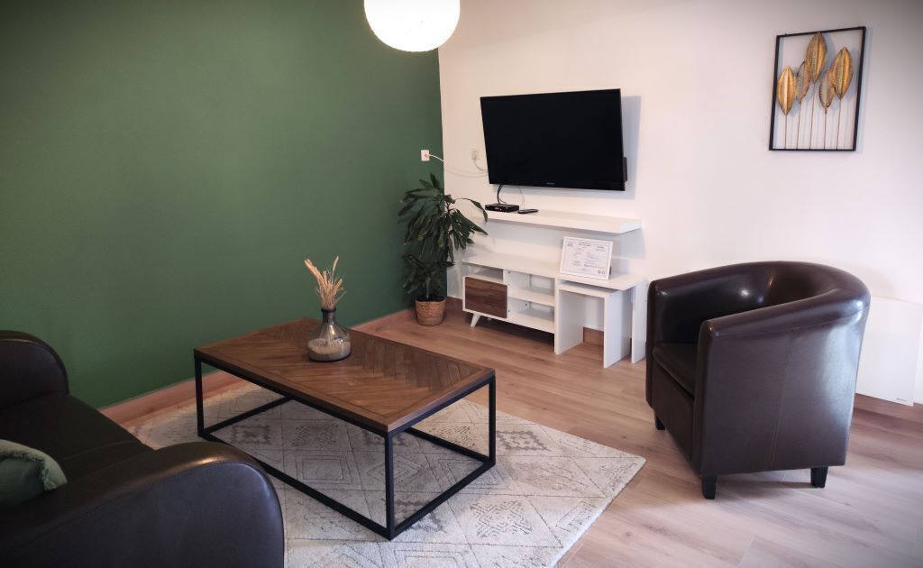 A modern living room with a green accent wall, a flat-screen TV mounted above a white console, a dark leather armchair, and a wooden coffee table with a vase.
