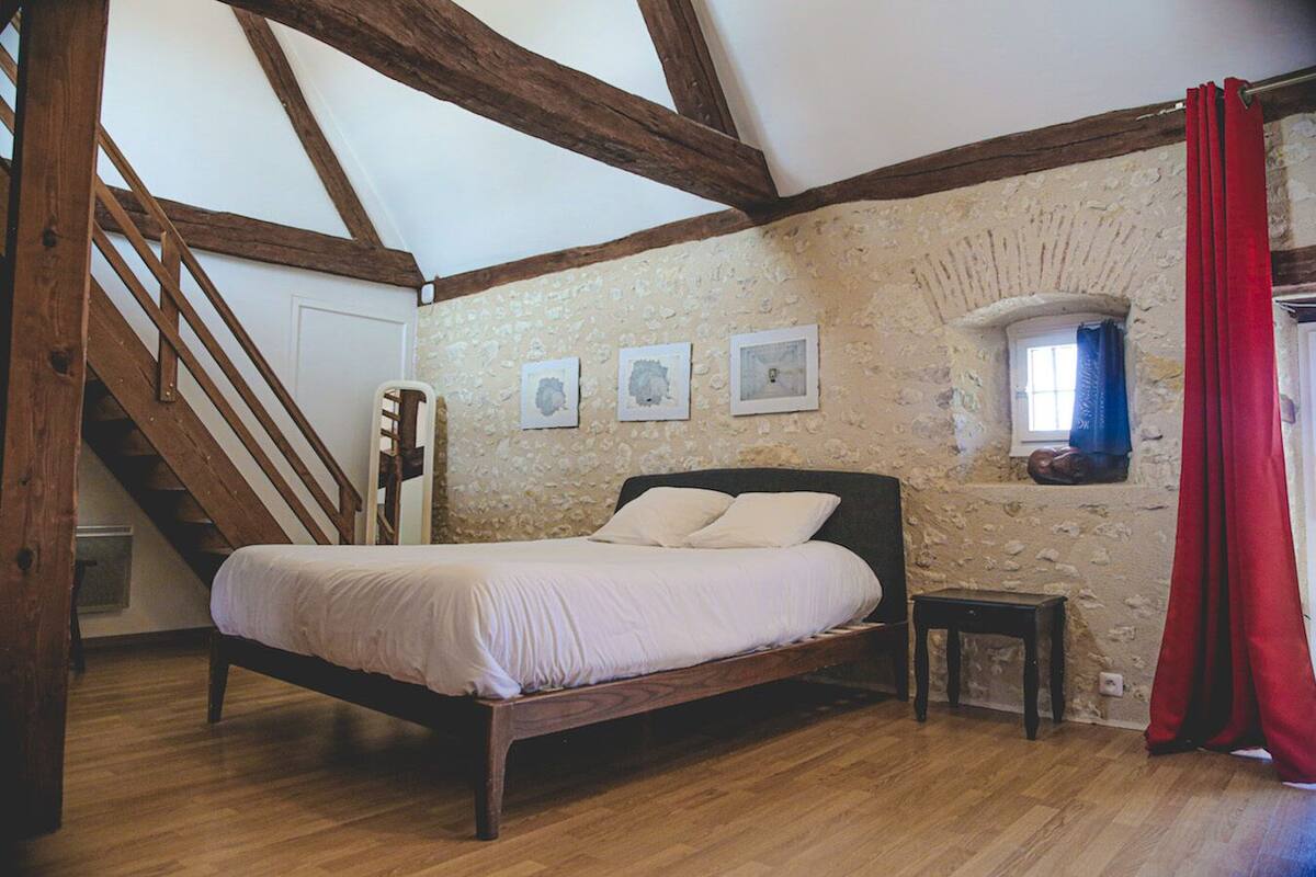 A bedroom with a wooden bed, a stone wall, a small window with a curtain, and a staircase.