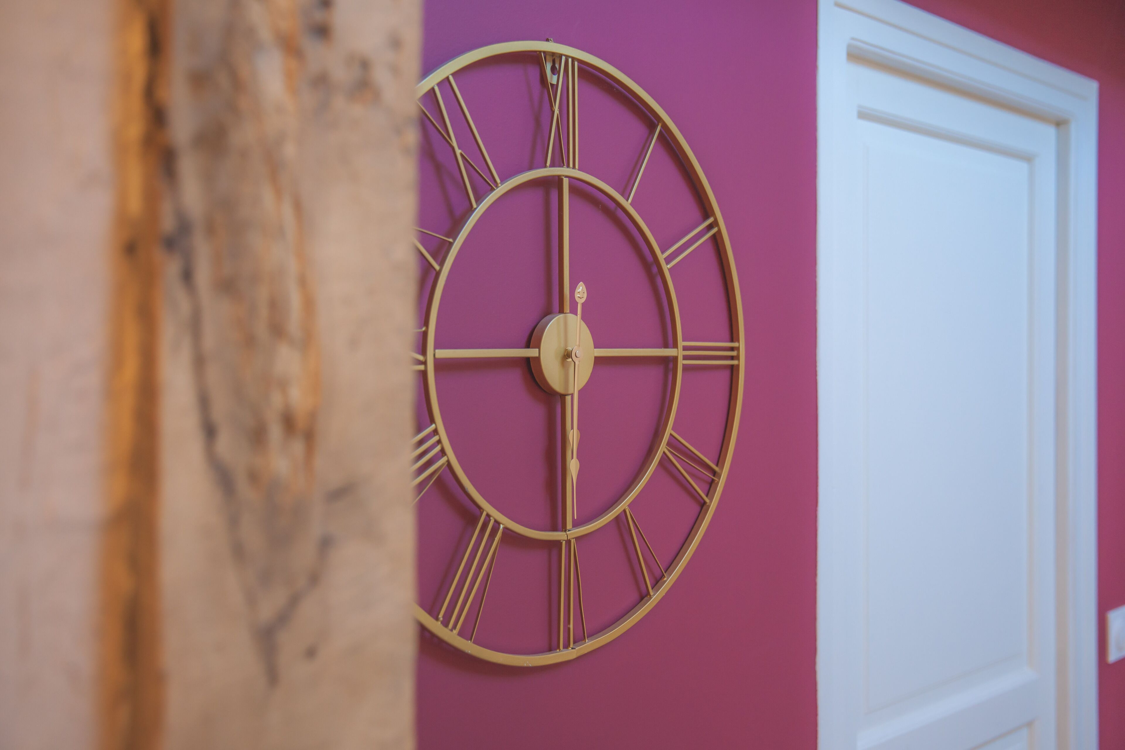 A wall-mounted clock with Roman numerals against a pink wall, next to a white door.