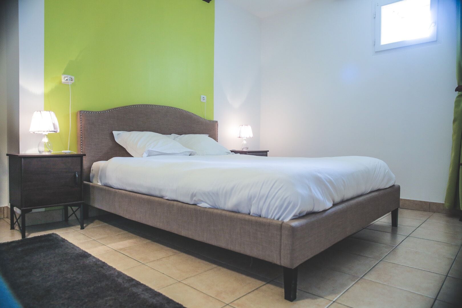 Un lit bien fait, avec du linge de lit blanc, deux tables de chevet avec des lampes, un mur d’accent vert et un sol carrelé.