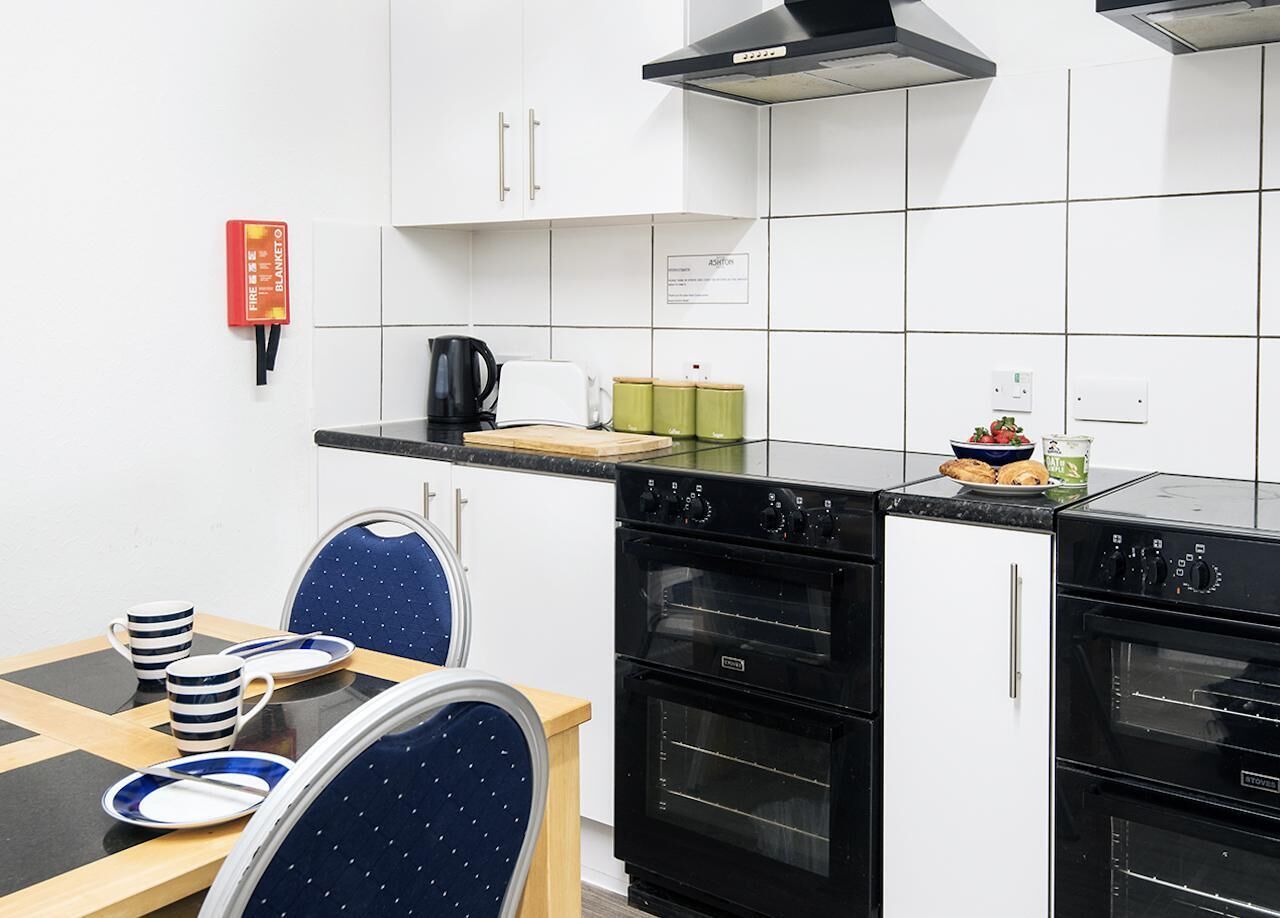 Una cocina compacta con armarios blancos, una estufa negra y un comedor con una silla azul y tazas a rayas.