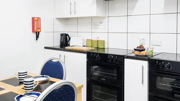 Una cocina compacta con armarios blancos, una estufa negra y un comedor con una silla azul y tazas a rayas.