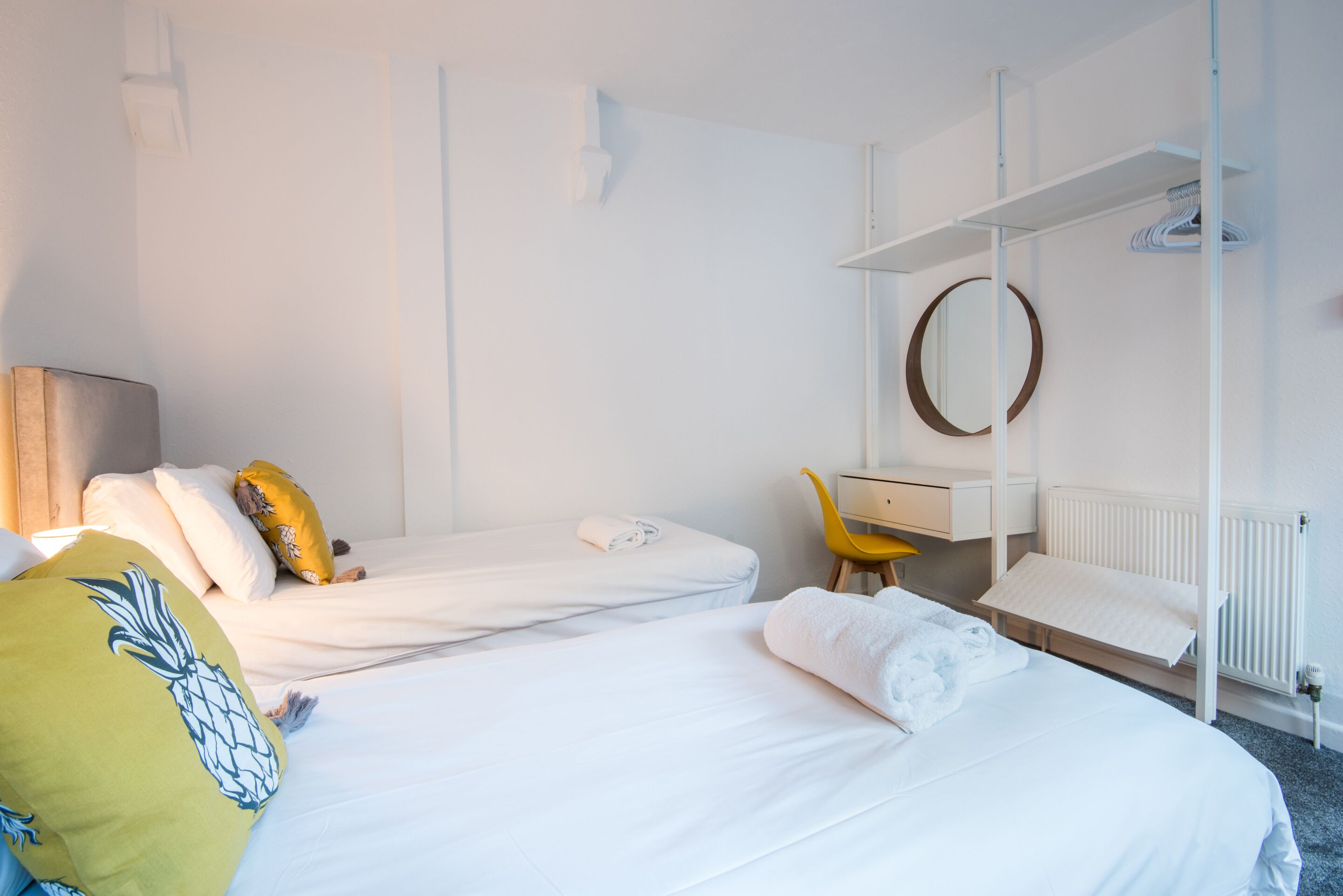 A hotel room with two beds, a desk, a chair, a round mirror, and a radiator.