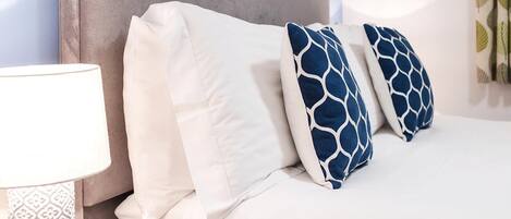 Un lit bien fait, avec une literie blanche et deux coussins bleus à motifs, une table de chevet avec une lampe à motifs et une tête de lit rembourrée de couleur grise.