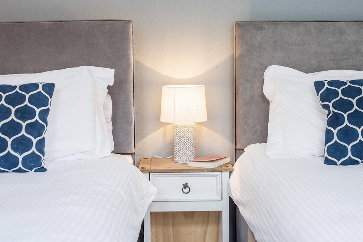 A double bed with white bedding and blue patterned pillows, a bedside table with a lamp, and a small drawer.