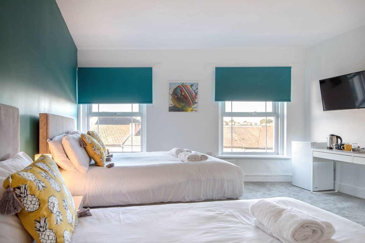 A hotel room with two beds, a desk, a television, and large windows with blue blinds.