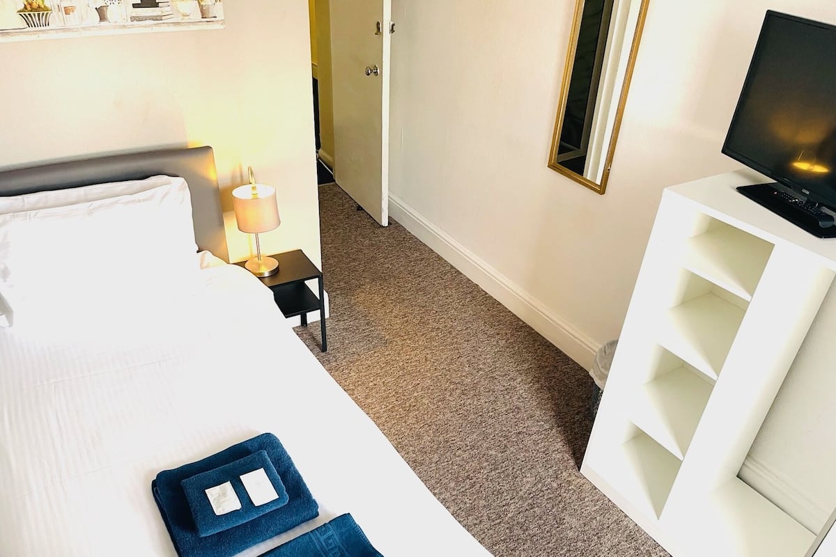 A hotel room with a bed, a TV on a stand, a small table with a lamp, and a mirror on the wall.