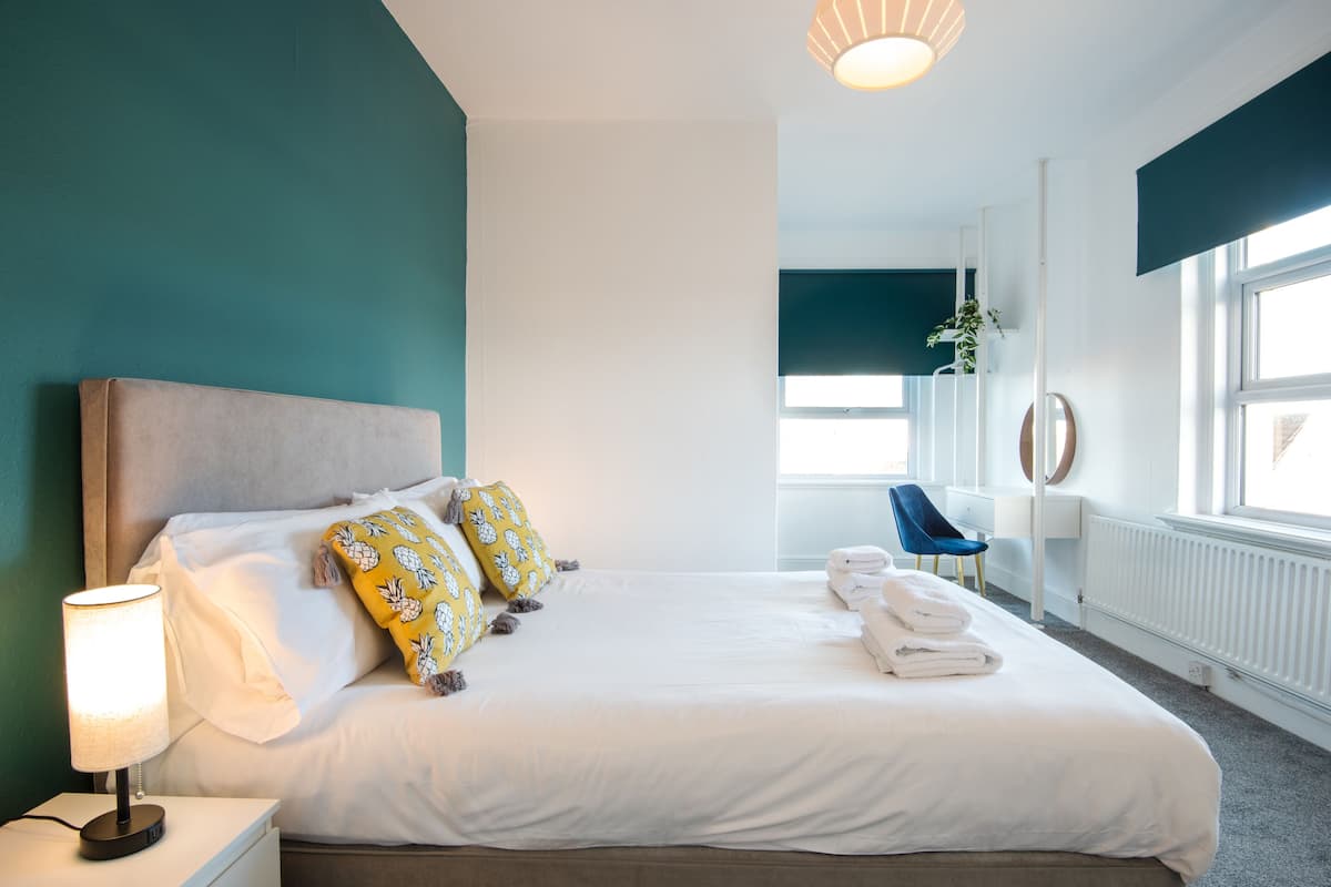 A neatly made bed with white linens and two decorative pillows, a bedside lamp, and a chair in a modern hotel room with teal walls and a large window.