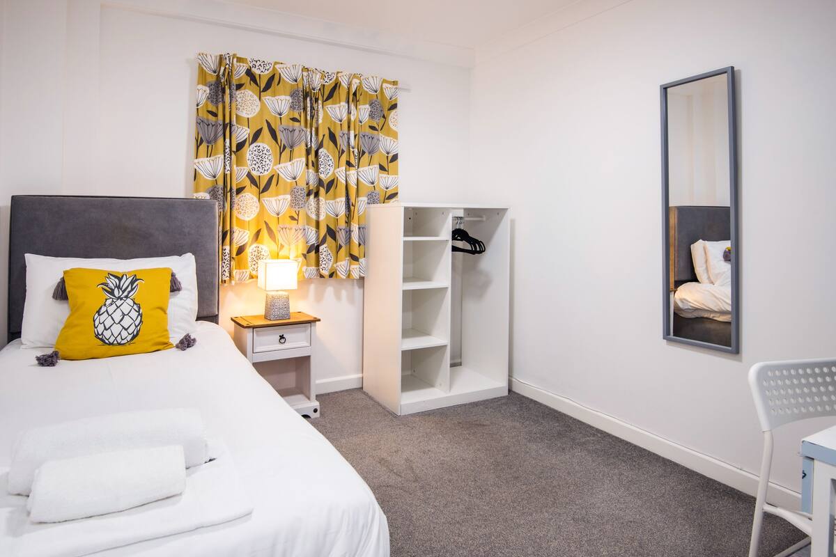A hotel room with a bed, a nightstand with a lamp, a wardrobe, a mirror, and a chair.