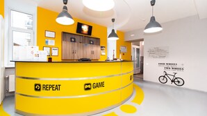 Ein Empfangsschalter mit Gelbtönen, ein Schild mit der Aufschrift 'REPEAT GAME' und eine Wand mit einer Fahrradillustration und motivierenden Text.
