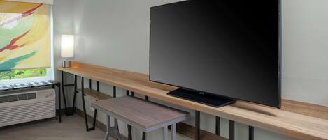 A modern room with a large flat-screen TV mounted on a wooden wall, a small stool, and a window with a colorful abstract painting.