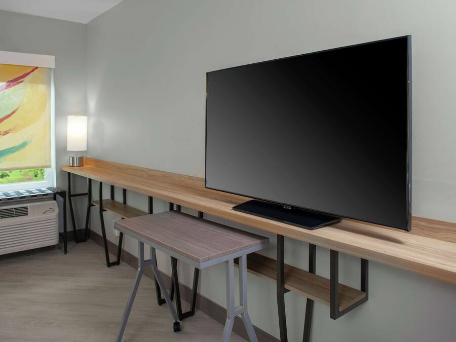 Una habitación moderna con una amplia televisión de pantalla plana montada en una pared de madera, un taburete pequeño y una ventana con un cuadro abstracto y colorido.
