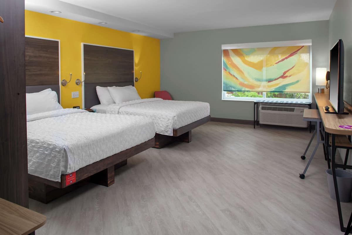 A hotel room with two beds, a desk, a chair, an air conditioning unit, and a large window with a view.