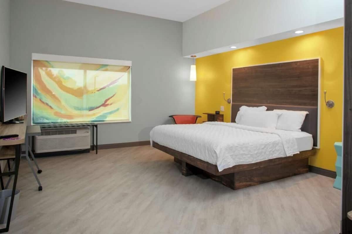 A bedroom with a bed, a desk, a chair, a TV, and a large abstract painting on the wall.