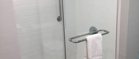 Ducha con cerramiento de vidrio, pared de azulejos blancos, puerta de cristal y toallero metálico con una toalla blanca.
