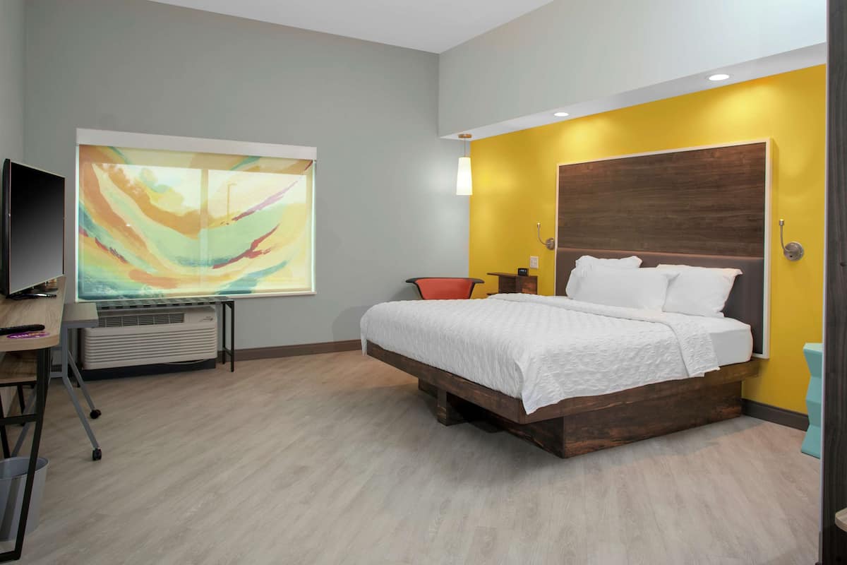 A modern bedroom with a wooden bed frame, a large abstract painting on the wall, and a flat-screen TV on a desk.