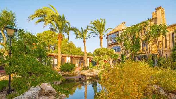 A serene courtyard with a small pond, palm trees, and a stone building with arched windows. - Villa Portol (Marratxi)