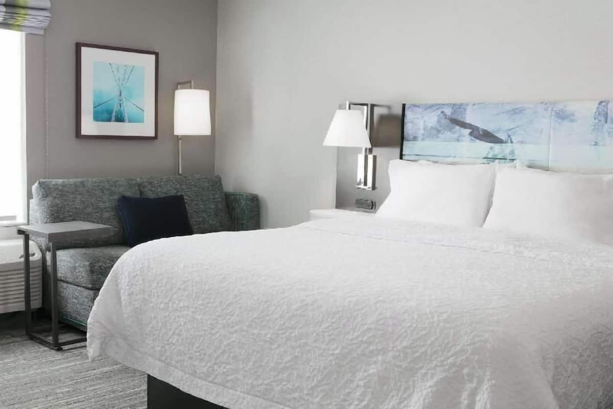 Une chambre d’hôtel avec un grand lit, un fauteuil gris, une table d’appoint, une lampe et deux œuvres d’art encadrées accrochées au mur.