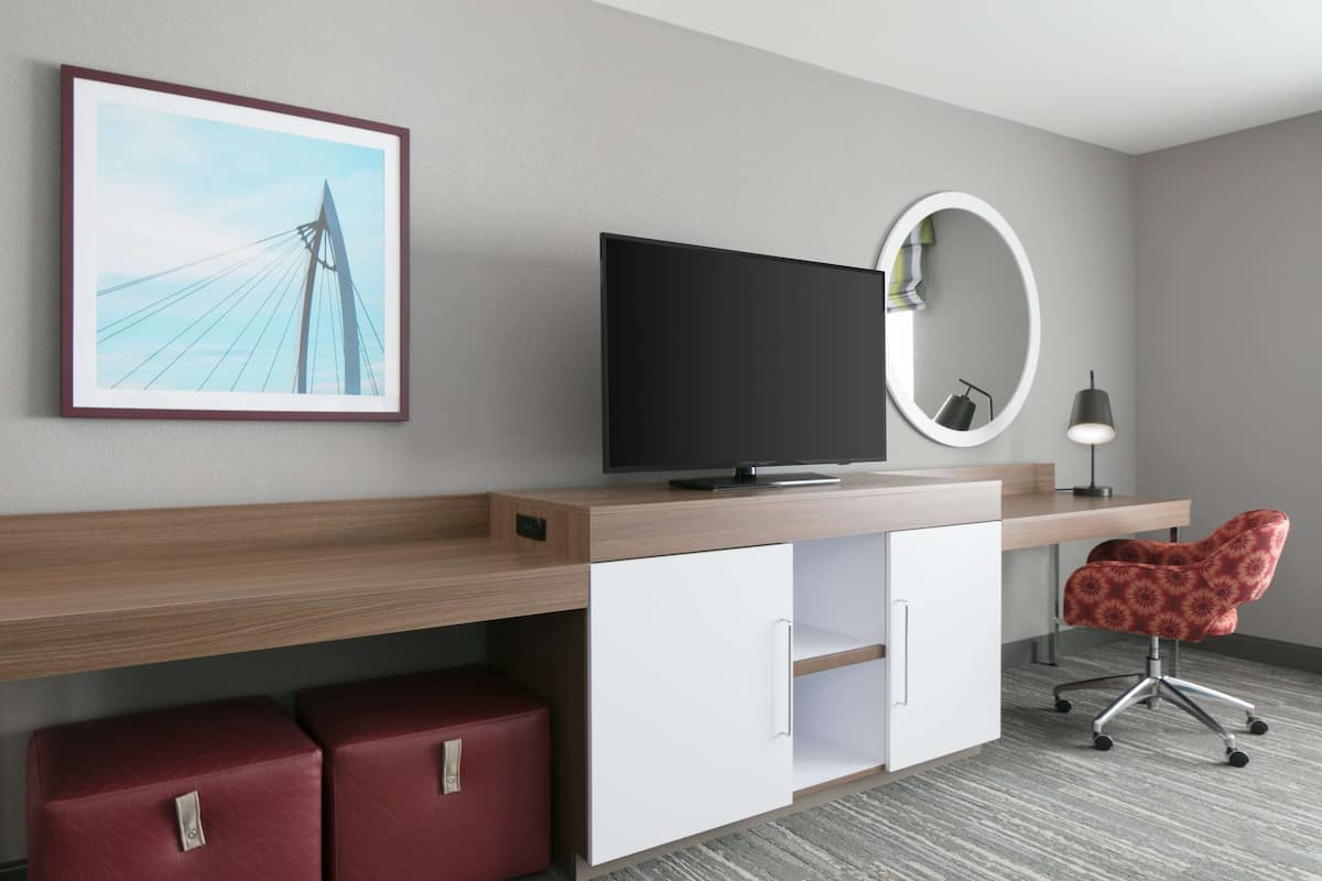 Une chambre d’hôtel moderne avec un bureau en bois, un téléviseur à écran plat, un fauteuil rouge rembourré, un miroir rond et un tableau encadré représentant un pont.